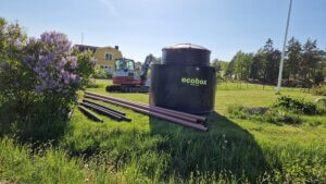 En stor, svart cylindrisk struktur märkt "ecobox small" ligger på en gräsyta med flera långa rör som ligger bredvid. I bakgrunden finns ett gult hus och några träd, med en klarblå himmel ovanför. Detta minireningsverk säkerställer effektiv vattenrening i kompakt form.